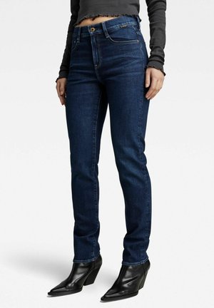Dunkelblaue Denim-Jeans mit Slim-Fit, hoher Taille, Fünf-Taschen-Design und dezentem Stick. Kombiniert mit schwarzen, knöchelhohen Stiefeln mit Absatz.