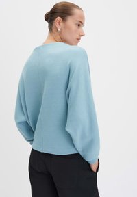 Pull léger bleu clair avec une coupe décontractée, présentant des manches larges, un détail de couture au dos et un col arrondi. Associé à un pantalon noir.