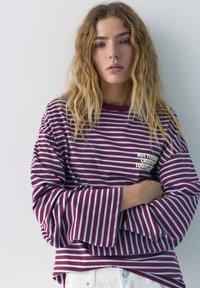 Jeune femme aux cheveux blonds ondulés portant un t-shirt à manches longues rayé bordeaux et bleu clair avec du texte sur la poitrine, debout devant un mur blanc.