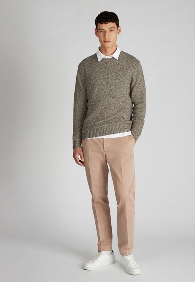 Ragazzo giovane in piedi che indossa un maglione di maglia marrone sopra una camicia bianca con colletto, pantaloni beige e sneakers bianche su uno sfondo uniforme.