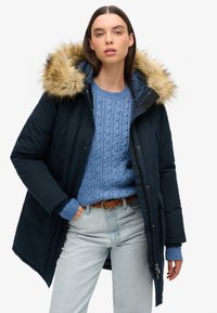 Parka navy avec capuche en fourrure, associé à un pull en maille torsadée bleu clair et à un jean en denim clair. Doté d'une fermeture éclair et d'une fermeture à bouton.