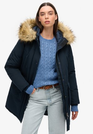 Parka azul marino con capucha forrada de piel, combinada con un suéter de punto en azul claro y jeans de mezclilla claros. Presenta un cierre de cremallera y botón.