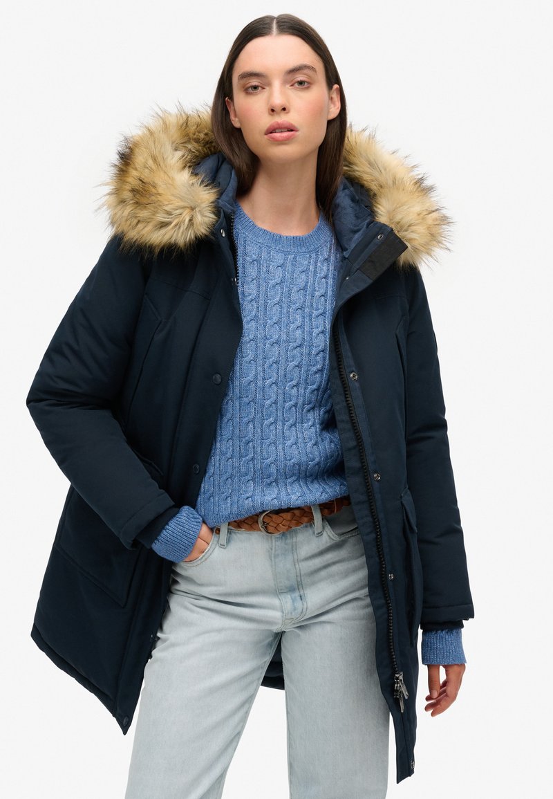 Parka navy avec capuche en fourrure, associé à un pull en maille torsadée bleu clair et à un jean en denim clair. Doté d'une fermeture éclair et d'une fermeture à bouton.
