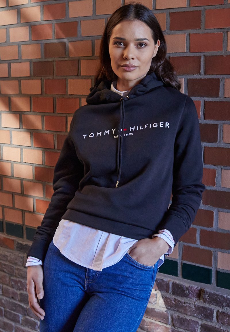 Čierna mikina s kapucňou s červeným a bielym logom "TOMMY HILFIGER" a "EST. 1985," nosená cez biele tričko, skombinovaná s modrými džínami.