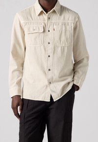Kremowa koszula z długim rękawem, posiadająca dwie przednie kieszenie, zapięcie na guziki i gładką fakturę. Stylizowana z ciemnymi spodniami cargo.