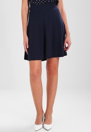 Jupe évasée bleu marine en tissu extensible, dotée d'une taille haute, atteignant le mi-thigh, assortie à des chaussures à talons hauts blanches.