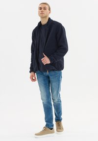 Laivastonsininen fleece-takki, jossa on korkea kaulus ja vetoketjukiinnitys, yhdistetty tumman yläosan, sinisten farkkujen ja vaaleanbeigejen kenkien, joissa on valkoiset pohjat kanssa.