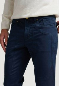 Pantalones de mezclilla azul marino con un corte ajustado, bolsillos estructurados y cierre de botón dorado; textura suave con detalles de costura sutiles.