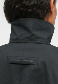 Chaqueta negra con un alto cuello, que presenta una textura suave y costuras visibles. Una etiqueta rectangular con "SAMSØE SAMSØE" está cosida en la parte posterior.