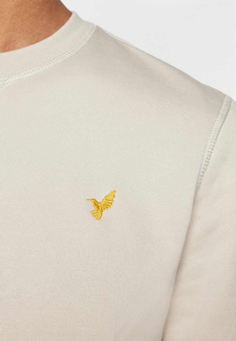 Sweatshirt léger beige clair à col rond en tissu doux, avec un petit logo d'oiseau jaune brodé sur la poitrine gauche.