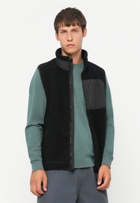 Zwarte fleece vest met hoge kraag en ritsclosure, met een voorkantzak met rits. De binnenlaag heeft een textuur. Licht blauw-groen shirt eronder.