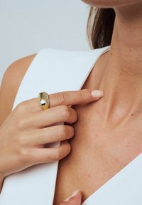 Guld ring med slät, böjd yta och texturerade detaljer. Finger som vilar mot huden nära nyckelbenet, med ljus nagellack.