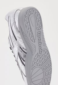 Srebrni tenisiki z razpokanimi vzorci, črnimi detajli in sivo gumijasto podplatom z teksturiranimi vzorci ter blagovno znamko.