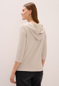 Kvinna med rakt ljusbrunt hår som bär en beige ribbad hoodie med trekvartsärmar och svarta jeans, vänd åt vänster.