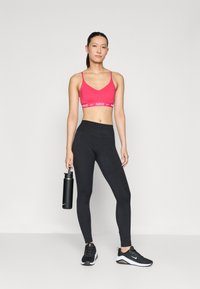Rozā sporta krūšturis ar balto Nike logotipu, melni augsti jostaslegu legingi un melni sporta apavi. Modelis tur melnu ūdens pudeli.