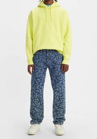 Sudadera con capucha de color amarillo neón con bolsillo frontal, combinada con jeans azules que presentan un patrón abstracto en gris. El calzado son zapatillas blancas.