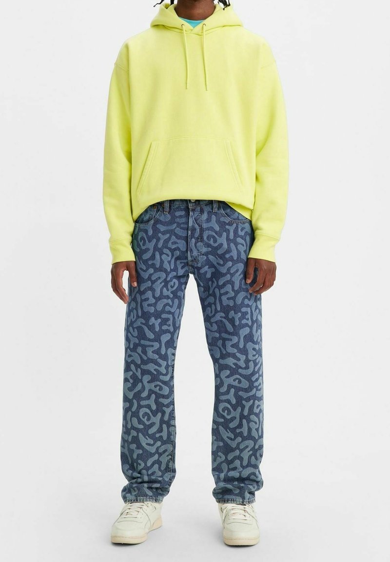 Sudadera con capucha de color amarillo neón con bolsillo frontal, combinada con jeans azules que presentan un patrón abstracto en gris. El calzado son zapatillas blancas.