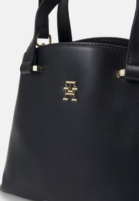 Sac à main en cuir noir avec logo en métal doré "TH" et poignées noires sur un fond clair uni.