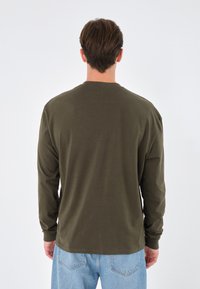 Langärmliges Shirt in dunklem Olivgrün, aus weichem Stoff gefertigt. Verfügt über einen Rundhalsausschnitt und eine lockere Passform mit gerippten Bündchen und Säumen.