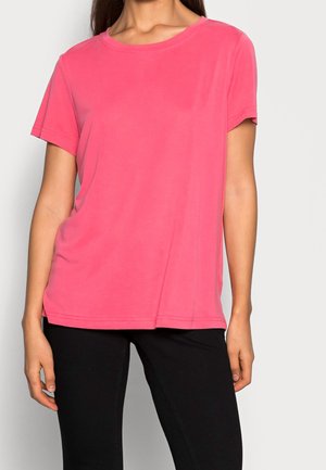 Woman wearing a bright pink short-sleeve t-shirt and black pants against a plain light background.