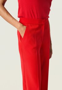 Červené kalhoty s hladkým povrchem, s širokým pasem a bočními kapsami. Střih je přiléhavý a rovný, zdůrazňující elegantní siluetu.