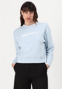 Világoskék pulóver bordázott mandzsettákkal és alsó résszel, melynek mellkasán fehér színű felirat olvasható: "KARL LAGERFELD JEANS". Puha, texturált anyag.
