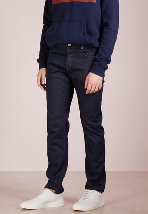 Person iført mørkeblå jeans, mørkeblå sweatshirt og hvide sneakers, stående mod en ensfarvet lys baggrund.