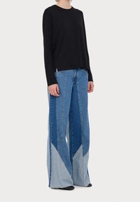 Haut noir à manches longues associé à un jean à jambes larges en denim à blocs de couleurs dans différentes nuances de bleu, présentant des motifs angulaires distincts.