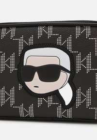 Mustat tekstuuripintaiset laukut, joissa on sarjakuvahahmo valkoisilla hiuksilla ja aurinkolaseilla, korostettuna valkoisilla pisteillä ja minimalistisilla suunnittelu-elementeillä.