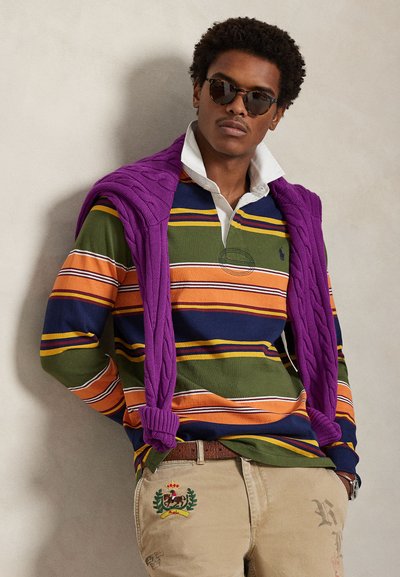 Ρουά πουκάμισο ράγκμπι με πολλαπλές ρίγες σε πράσινο, μπλε, πορτοκαλί και λευκό χρώμα· φορεμένο πάνω από μοβ πουλόβερ με πλέξη καλωδίου. Τα μπεζ παντελόνια έχουν κεντημένο σχέδιο.