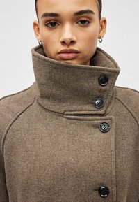 Pessoa a usar um casaco castanho com gola alta, botões pretos, brincos prateados em forma de argola e expressão facial neutra, contra um fundo branco.