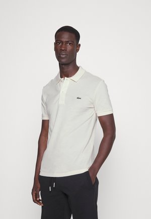 Creme polo shirt med korte ærmer, med krave og et grønt broderet logo på venstre bryst. Blødt bomuldsstof.