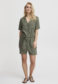 Olivgrüner Leinen-Jumpsuit mit kurzen Ärmeln, Knopfleiste, Brusttaschen und gebundenem Taillenbund. Getragen mit flachen Sandalen, vor einem schlichten Hintergrund.