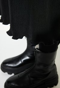 Zwart ribbelstof met gegolfde randen valt over elegante zwarte enkellaarsjes met een rits en een gestructureerde zool. Matte en glanzende contrasten zijn duidelijk zichtbaar.