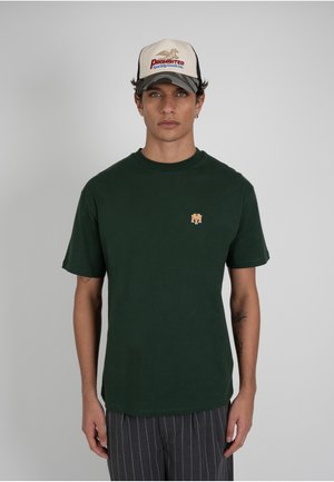 Nuori mies, jolla on päällään tummanvihreä t-paita, jossa pieni oranssi logo, sekä valkoinen ja harmaa baseball-lippis, jossa on teksti "Prohibited Sporting Goods Inc.".