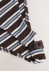 Gros plan d'une manche en tissu côtelé rayé marron, blanc et bleu avec des boutons-pression noirs sur fond beige.