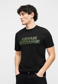 Άνδρας με σγουρά μαλλιά που φοράει μαύρο T-shirt με πράσινο κείμενο «ARMANI EXCHANGE», στέκεται με τα χέρια στις τσέπες, κοιτάζοντας αριστερά σε απλό φόντο.