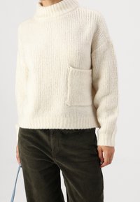 Pull en grosse maille crème avec un col roulé, une texture côtelée et une poche avant. Associé à un pantalon en velours côtelé vert foncé et une sangle bleue.