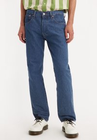 Blå denimjeans med rak bendesign, med fem fickor och synliga sömmar. Bärs med vita skor och en grönrandig skjorta.