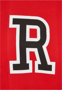 Rød stoff med et stort svart "R" innrammet i hvitt. Teksturen ser glatt ut, og designet er sentrert på plagget.