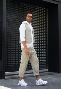 Colete cinzento claro sobre um hoodie branco, combinado com calças cargo claras e ténis brancos. Tecido texturizado e vários bolsos são visíveis.