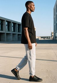 Mann läuft draußen und trägt ein schwarzes T-Shirt, hellgraue Jogginghose und schwarze Turnschuhe zwischen modernen Betongebäuden im Tageslicht.