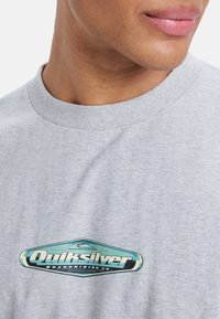Gray cotton t-shirt featuring a blue and black Quiksilver logo graphic on the chest, with a crew neckline and a standard fit.