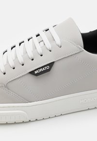Baskets grises en matériau synthétique lisse, avec des lacets blancs, une doublure intérieure noire et un patch logo noir sur le côté. Semelle blanche texturée.