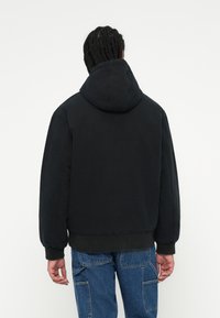 Person med fletninger iført en sort hættejakke og blå cargo jeans, vendt væk mod en ensfarvet hvid baggrund.