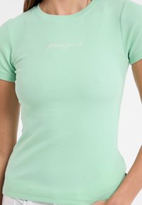 Mėtų žalios spalvos dryžuota marškinė su trumpomis rankovėmis ir apvaliu kaklu. Ant krūtinės yra balta siuvinėta "guessjeans" logotipas.