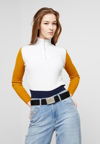 Ribbade tröja med halvdragkedja, i vitt, senapsgult och marinblått. Kombineras med blekta blå jeans och ett svart vävt bälte med metallspänne.