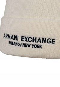 Cream-colored knit beanie with a textured surface, featuring black embroidered text: "ARMANI EXCHANGE MILANO / NEW YORK" along the brim.