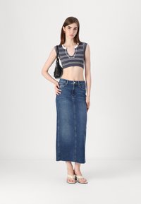 Cropped top s črno-belimi črtami in V-izrezom, v kombinaciji z dolgim jeans krilom. Model drži črno torbico in nosi bež sandale.
