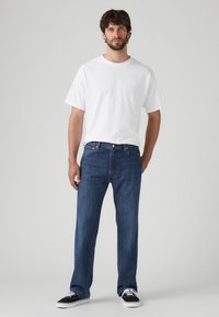 Camiseta de algodón blanca combinada con jeans de mezclilla de pierna recta azul. El modelo lleva zapatillas negras con suelas blancas, de pie frente a un fondo neutro.